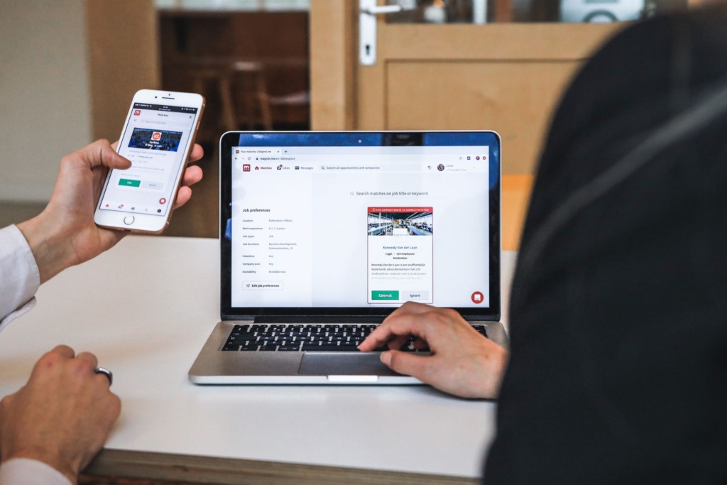 Person holding smartphone next to laptop displaying website interface at wooden desk